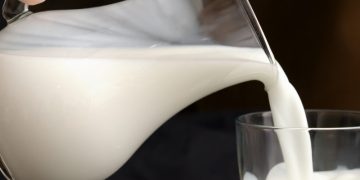 Woman pouring fresh milk from jug into glass on table